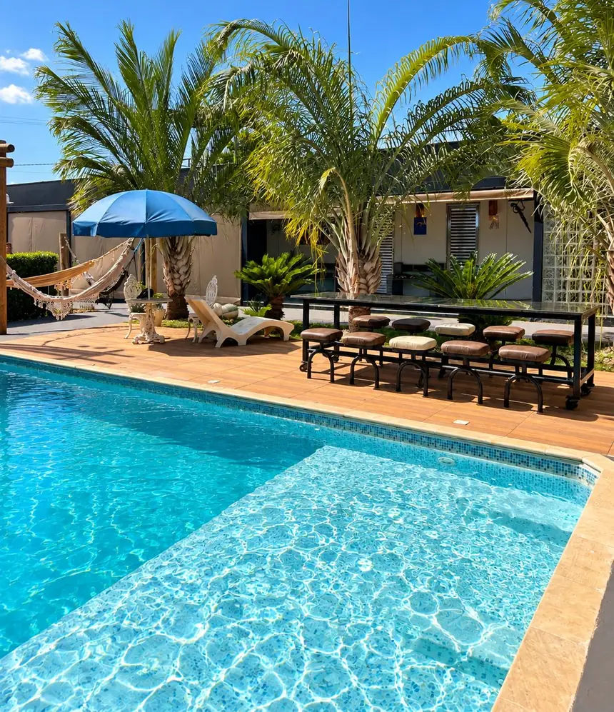 Piscina aquecida com deck de madeira, guarda-sol azul e rede entre as palmeiras